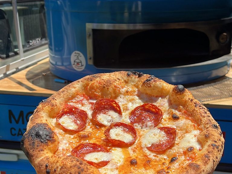 Wood-fired pepperoni pizza held up in front of the blue Antimo Pizza Chef truck, pizza catering Adelaide