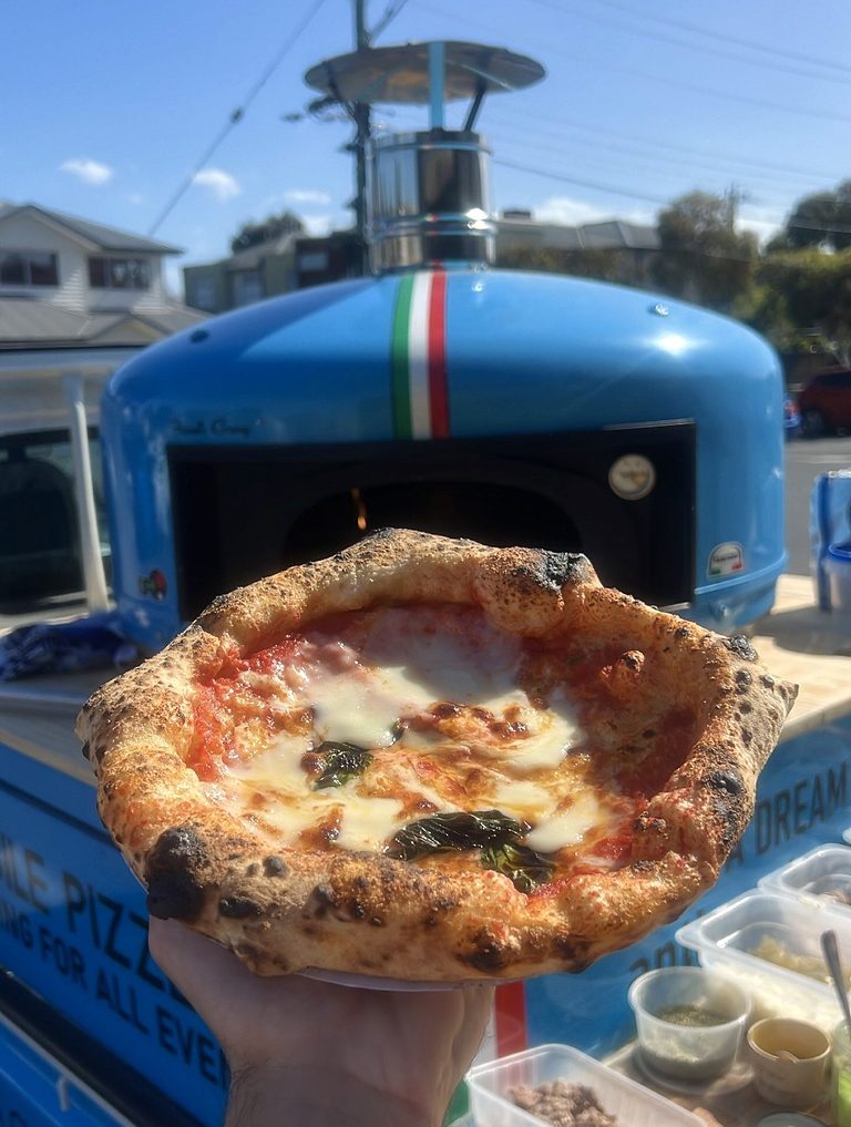 Wood-fired Margherita held up in front of the lit blue oven under a clear blue sky, pizza catering Adelaide CBD