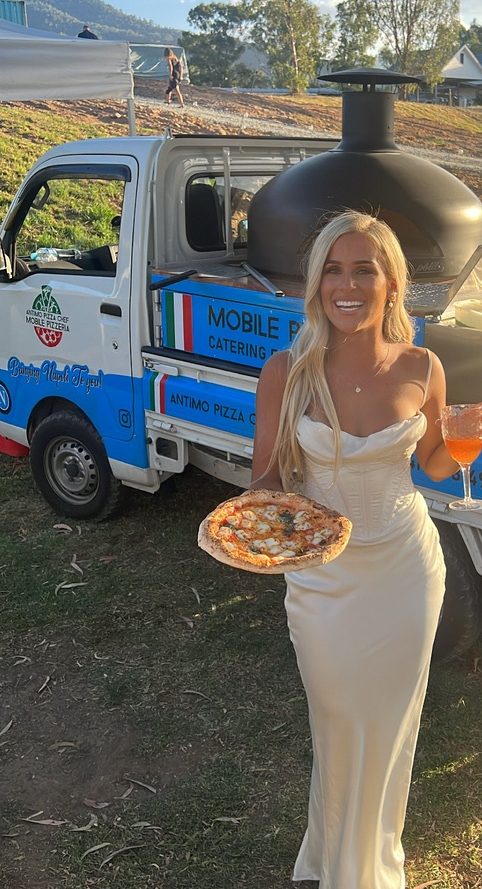 Bride in a white gown holding a wood-fired pizza and a spritz beside our pizza truck at sunset