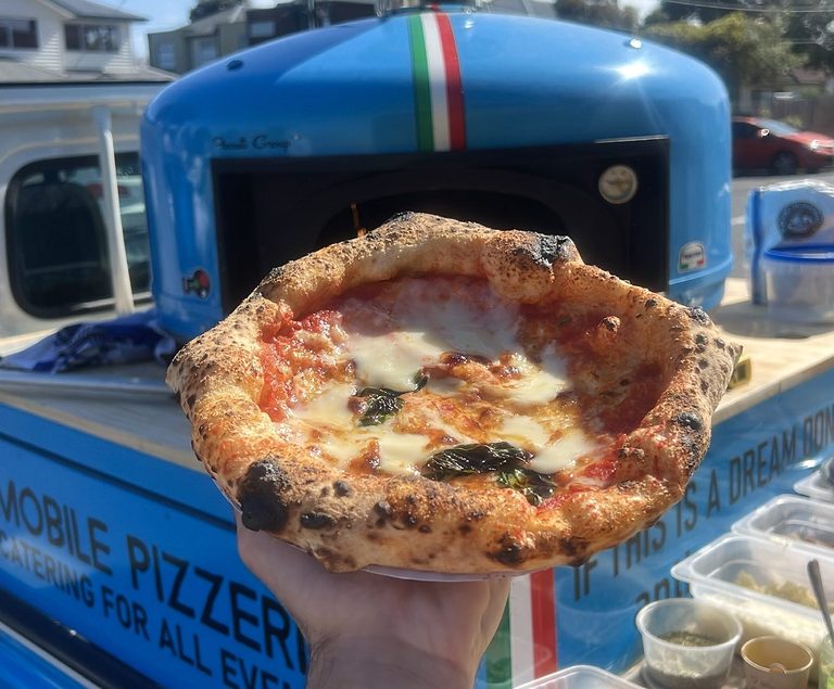 Wood-fired Margherita held up in front of the blue oven on our pizza truck