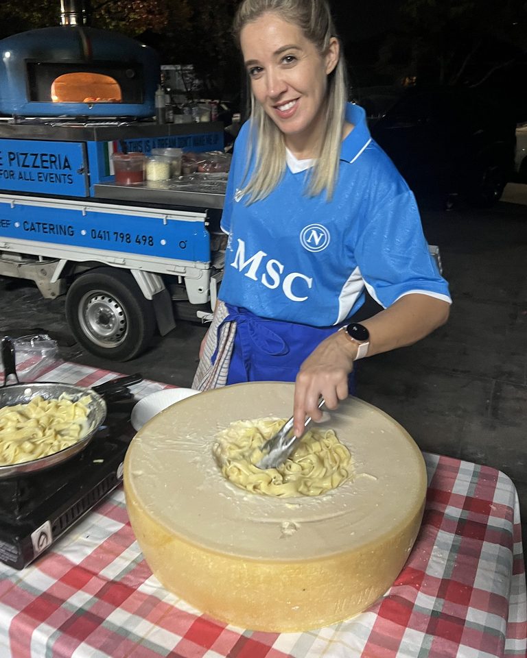 Our chef tossing fresh fettuccine pasta inside a giant hollowed-out Parmigiano wheel in front of the blue Antimo Pizza Chef truck