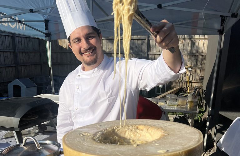 Chef lifting fresh spaghetti out of a giant cheese wheel at an outdoor event