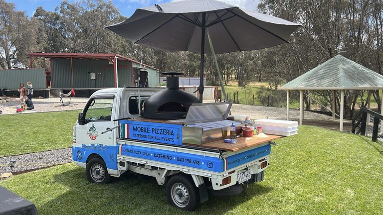 The blue and white pizza truck with its black wood-fired oven, set up on a lawn under an umbrella
