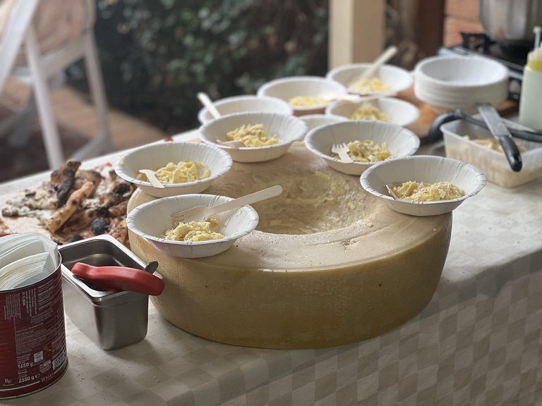 Hollowed-out Parmigiano Reggiano cheese wheel surrounded by bowls of fresh pasta at an outdoor event