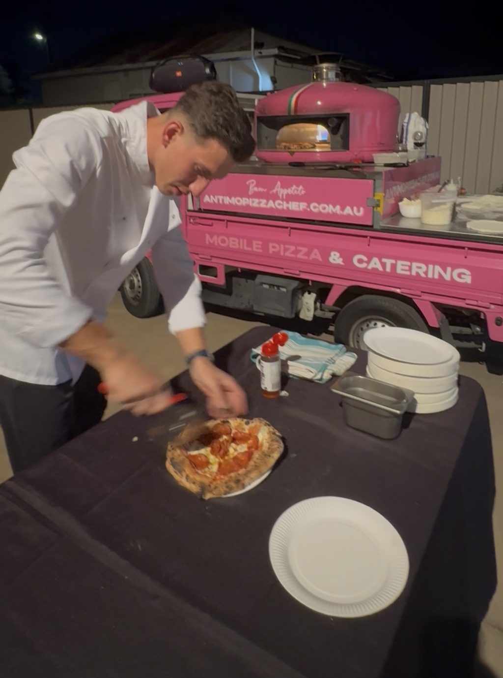Matteo slicing a fresh wood-fired pizza beside our pink truck at night