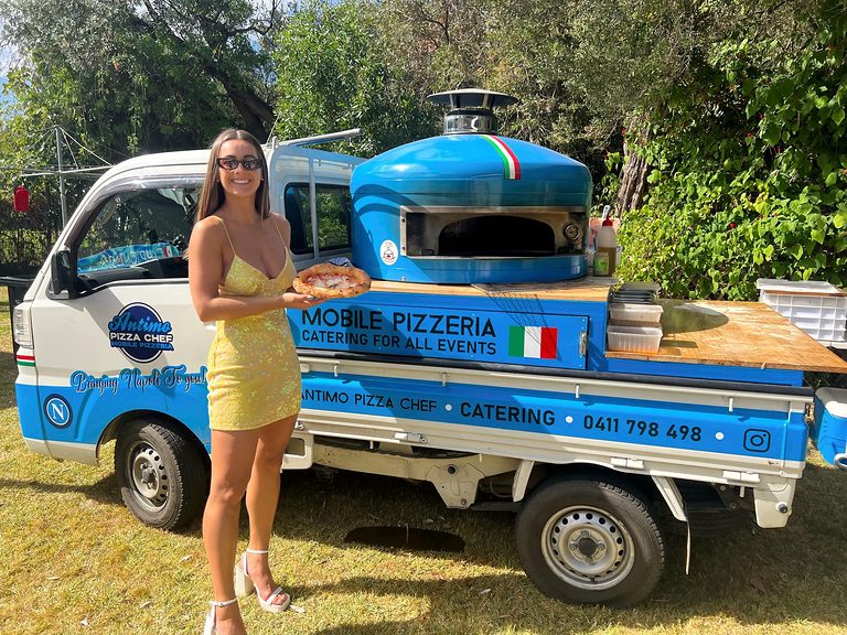 Guest holding a fresh wood-fired pizza in front of the blue Antimo Pizza Chef truck in Sydney