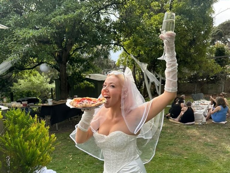 Bride in her wedding gown holding a fresh wood-fired pizza and raising a champagne glass